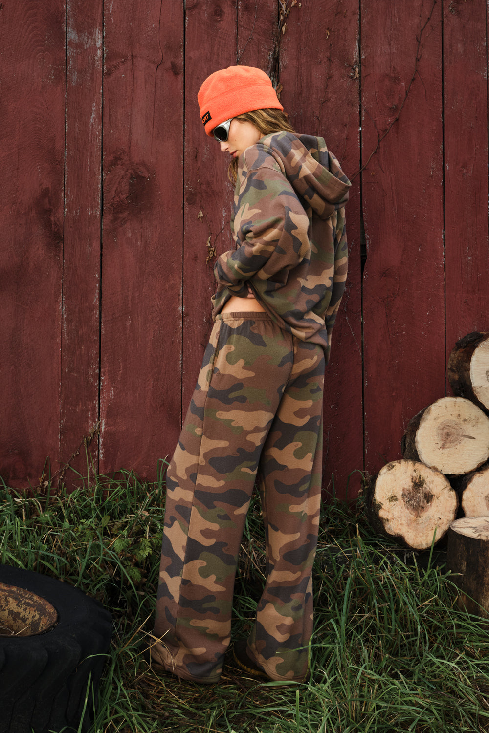 A person wearing a camouflage hoodie and matching wide-leg sweatpants stands outdoors in front of a red wooden fence, with grass and stacked wood logs visible.
|M1
