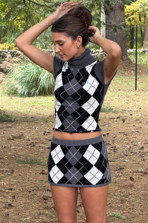 A woman wearing a matching two-piece argyle knit set consisting of a short sleeve turtle-neck crop top and a mini skirt, standing outdoors in a park-like setting.
|M2