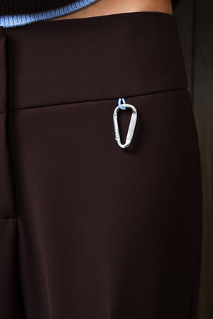 A close-up of dark brown trousers featuring a silver metal carabiner accessory attached to the side waistband.