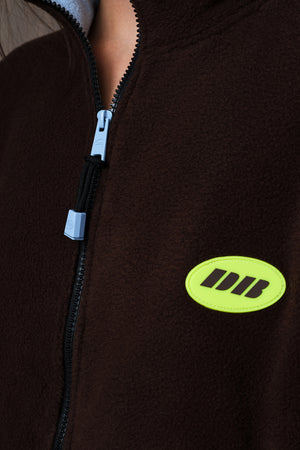 Close-up of a dark brown fleece zip-up jacket with a black zipper and a bright neon yellow oval logo patch featuring the letters 'DB'.