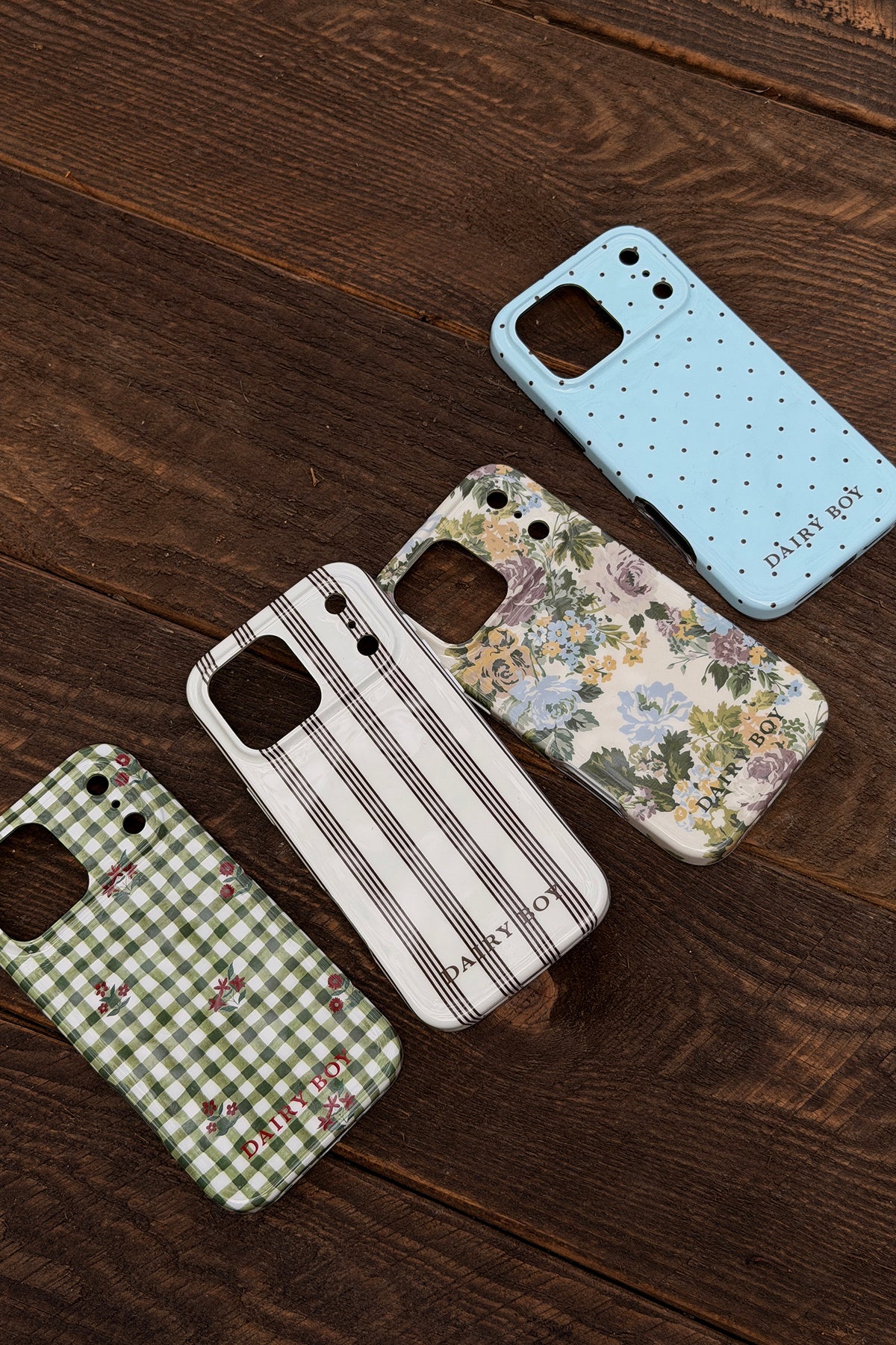 Light blue phone case with small dark polka dots and DAIRY BOY lettering, shown back-side up beside three other patterned cases.