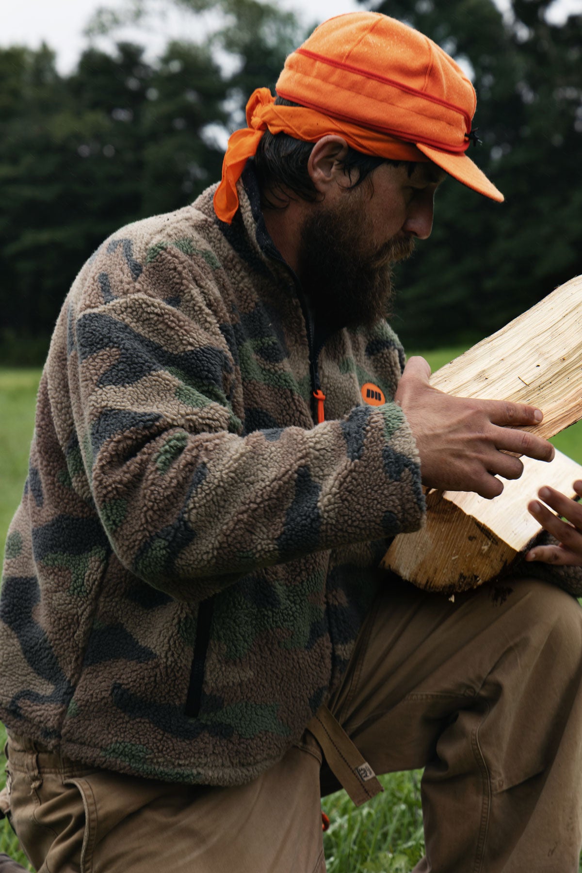 A bearded man wearing a brown camouflage fleece jacket, khaki pants, and an orange cap sits outdoors in a grassy field while holding a cut piece of wood.