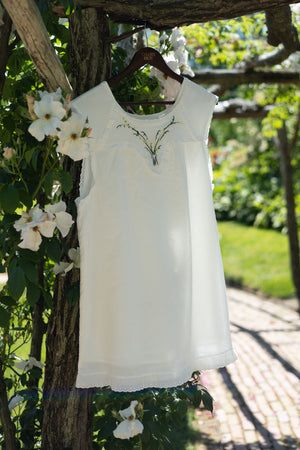 A white sleeveless dress with delicate floral embroidery on the chest, hanging on a wooden hanger from a rustic garden trellis.