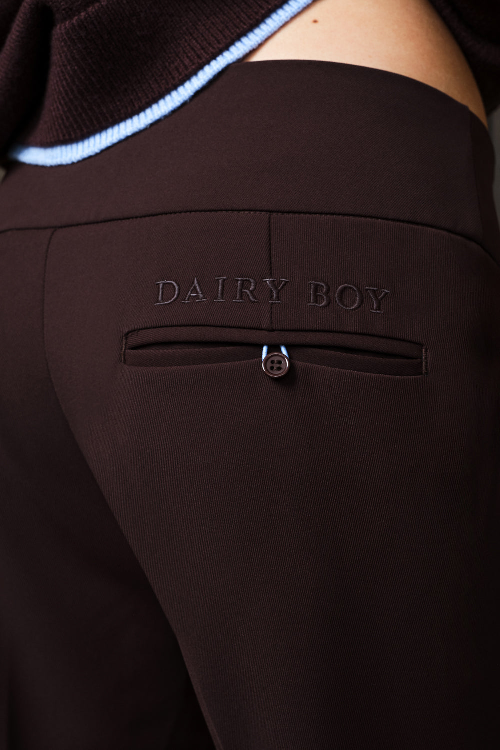 Close-up of dark brown pants with an embossed 'DAIRY BOY' logo on the back pocket, worn by a model with a light blue striped knit top visible at the top.
|M1