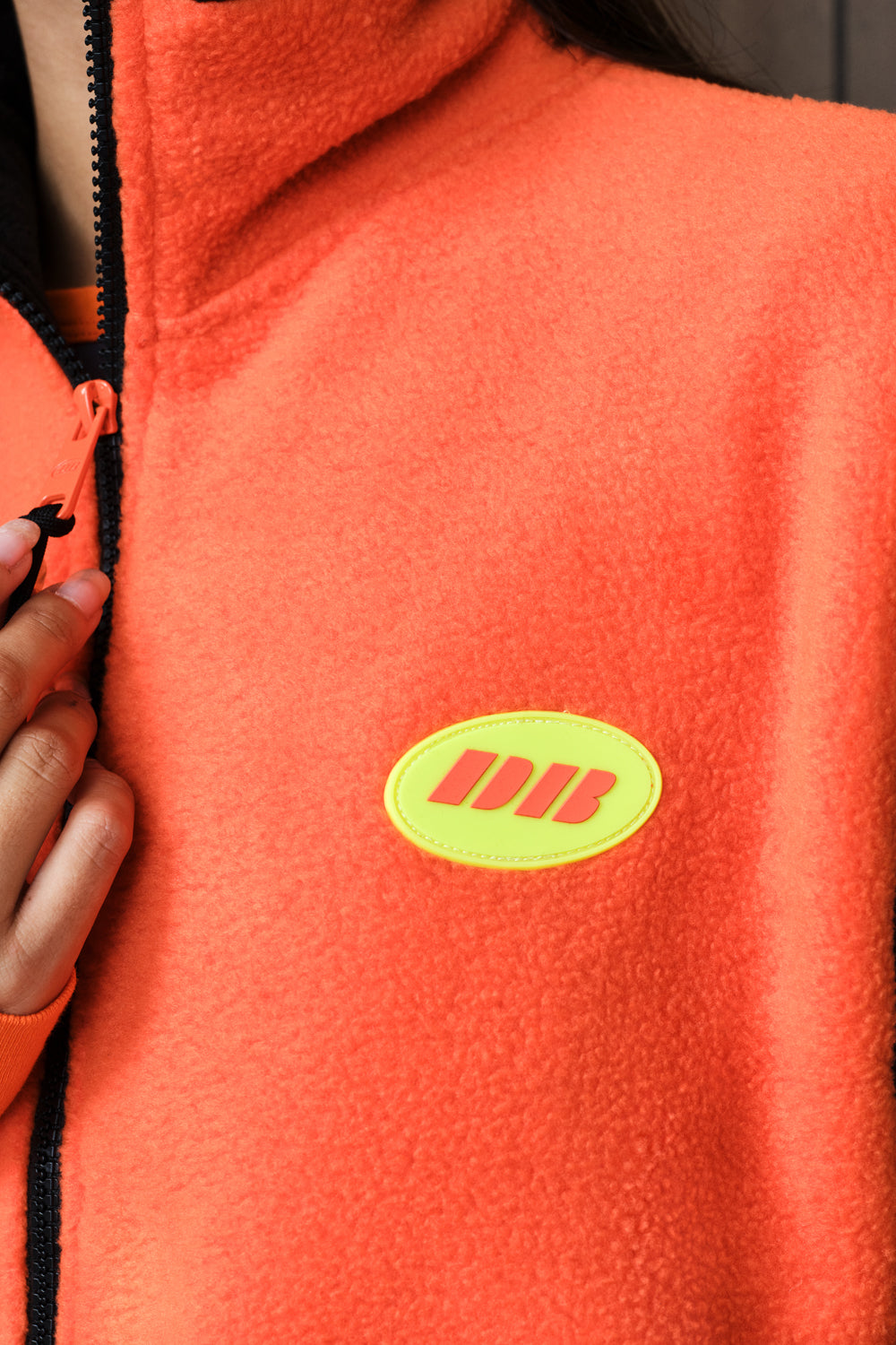 Close-up of a bright orange fleece zip-up vest with a black zipper and a yellow oval chest patch featuring yellow stylized lettering, worn by a person whose hand is visible at the zipper.