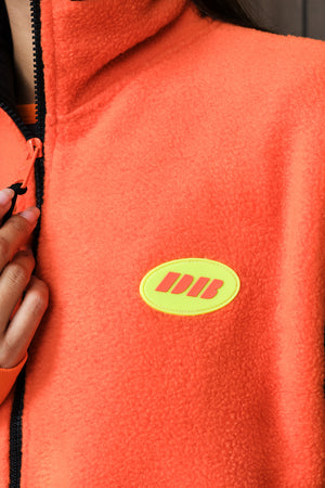 Close-up of a bright orange fleece zip-up vest with a black zipper and a yellow oval chest patch featuring yellow stylized lettering, worn by a person whose hand is visible at the zipper.