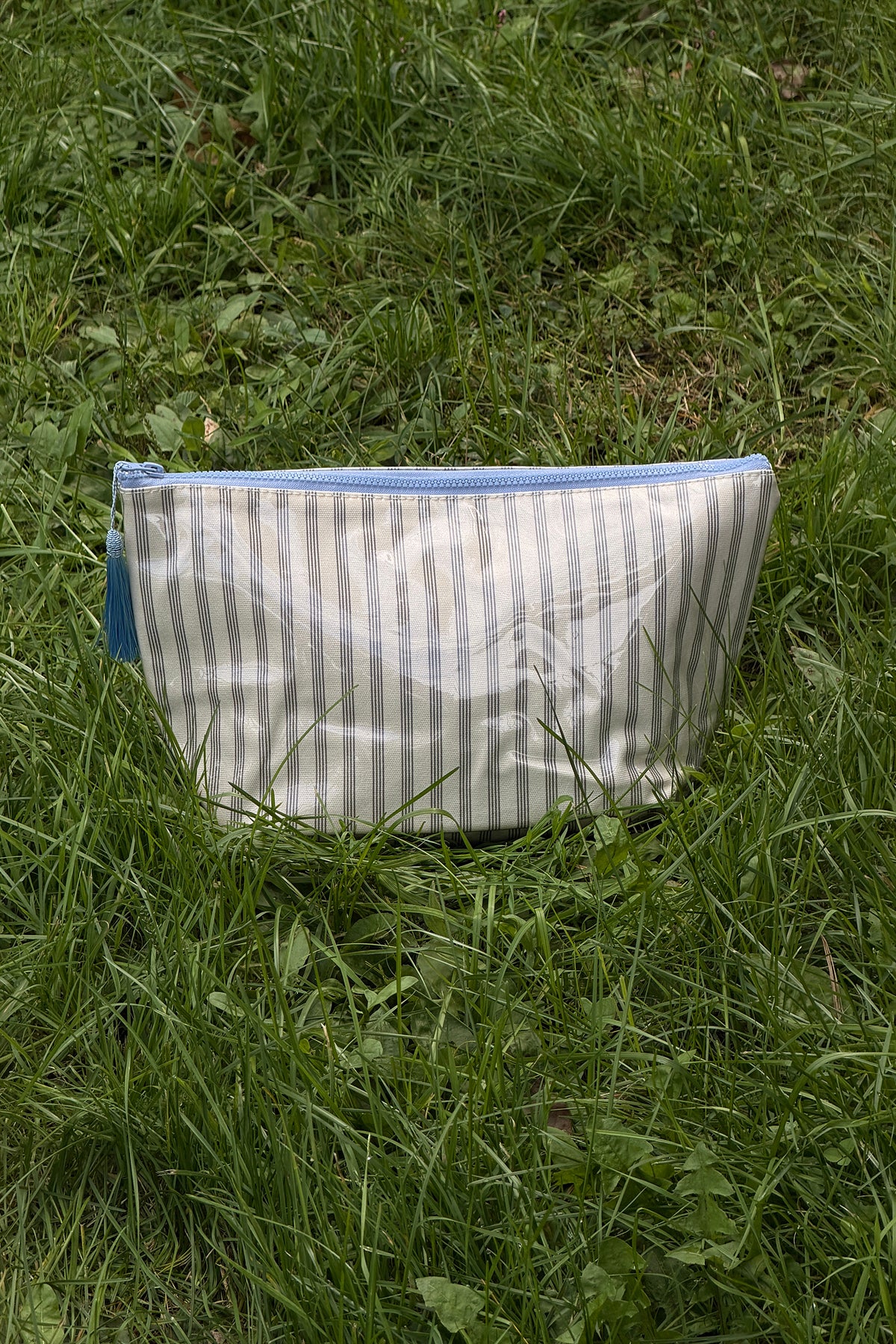 Front view of a cream makeup pouch with thin brown vertical stripes, a light blue zip along the top and a blue tassel pull.