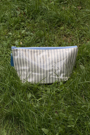 Front view of a cream makeup pouch with thin brown vertical stripes, a light blue zip along the top and a blue tassel pull.