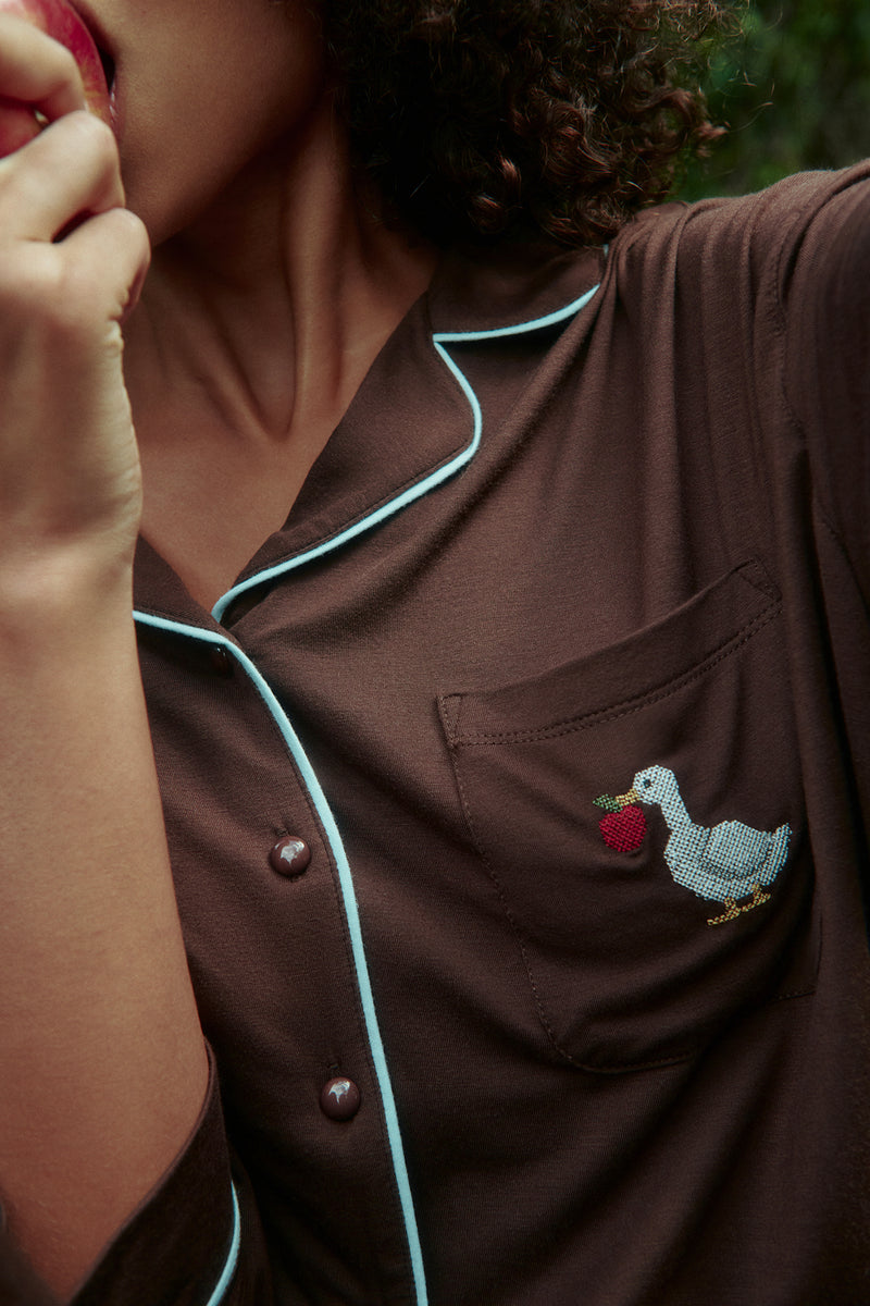 Detail of a brown pajama shirt with light blue piping, buttons and a chest pocket with a white goose and red apple motif.