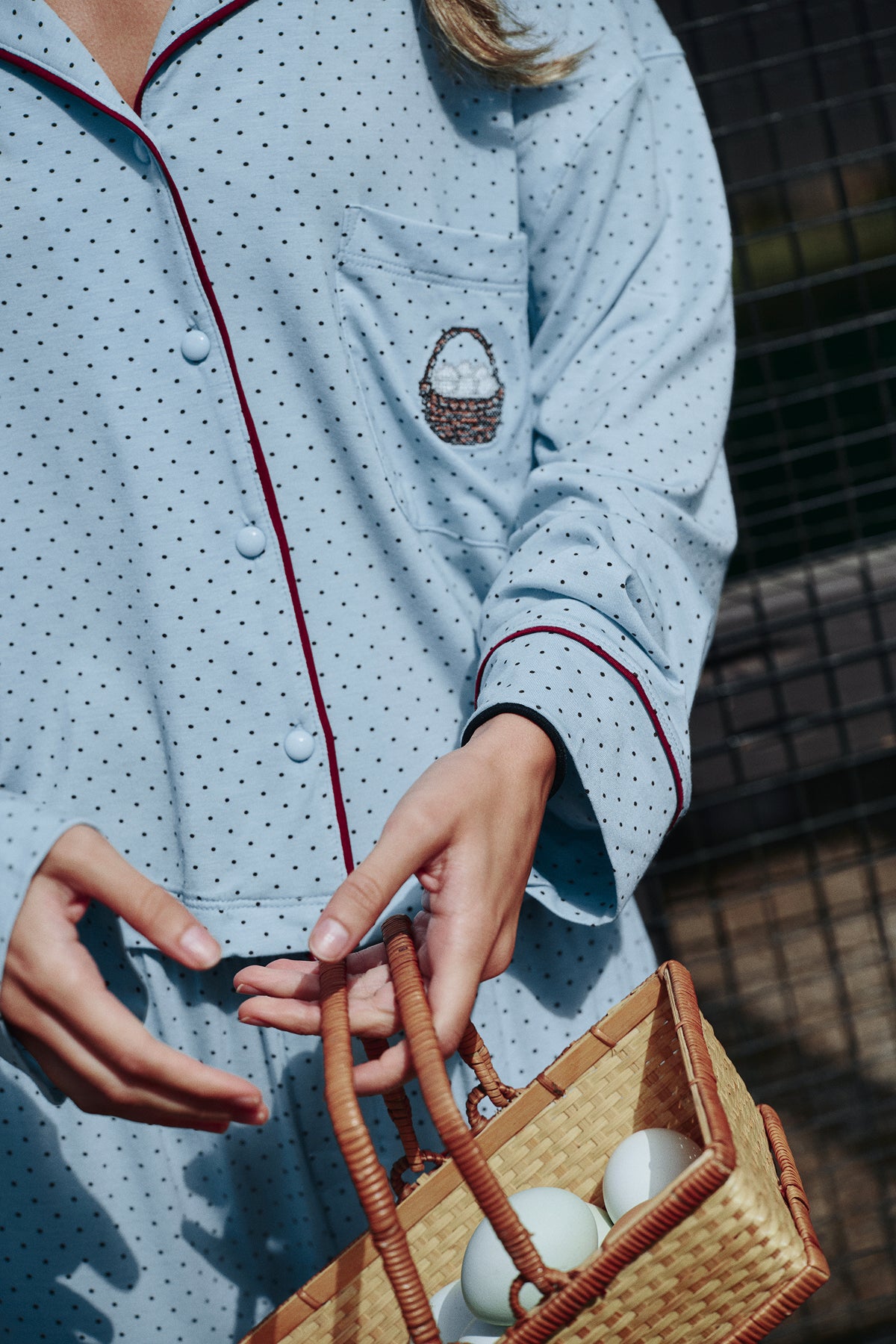 Detail of a light blue polka-dot pajama shirt with burgundy piping, covered buttons and a chest pocket with a small basket-of-eggs motif, on a model.