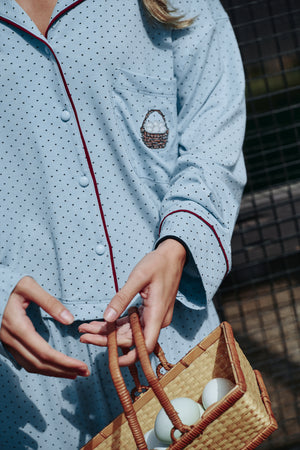 Detail of a light blue polka-dot pajama shirt with burgundy piping, covered buttons and a chest pocket with a small basket-of-eggs motif, on a model.