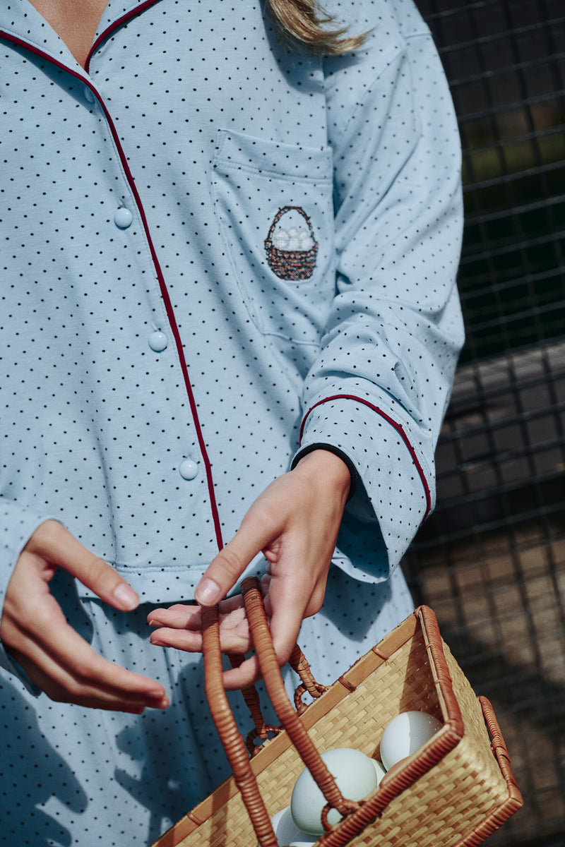 Detail of a light blue polka-dot pajama shirt with burgundy piping, covered buttons and a chest pocket with a small basket-of-eggs motif, on a model.