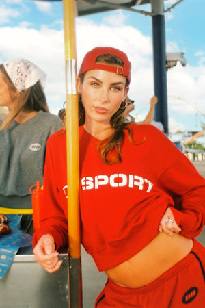 A woman wearing a red cropped sweatshirt with 'DB SPORT' printed in white across the chest, matching red sweatpants, and a red cap, standing outdoors near a pole.
|M2