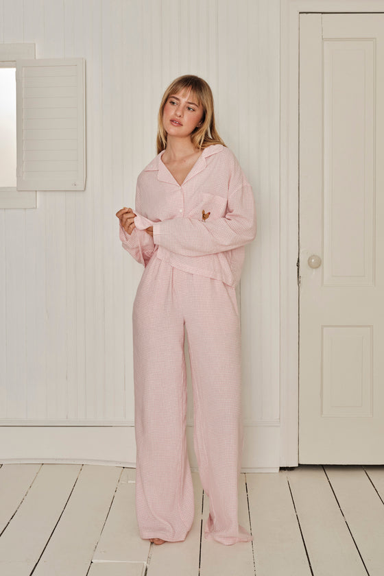 Woman wearing a pink gingham sleep set in a minimalistic room with white walls and wooden floor. |M1