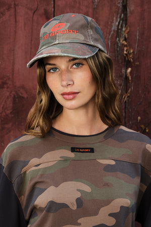A woman wearing a camouflage baseball cap and a matching camouflage crewneck shirt, posed against a rustic reddish-brown wooden background. The camo pattern features shades of green, brown, and beige, with orange-red embroidered 'DB SPORT' branding on the cap and a small black brand patch on the sweatshirt.