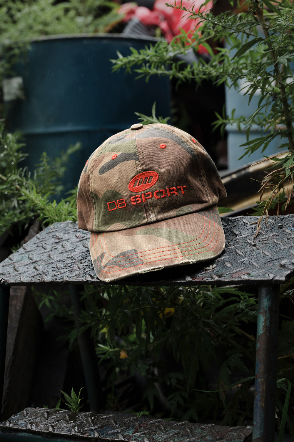 A camouflage baseball cap with red embroidered 'DB SPORT' text and logo, resting on a rusty metal table outdoors amidst green foliage.