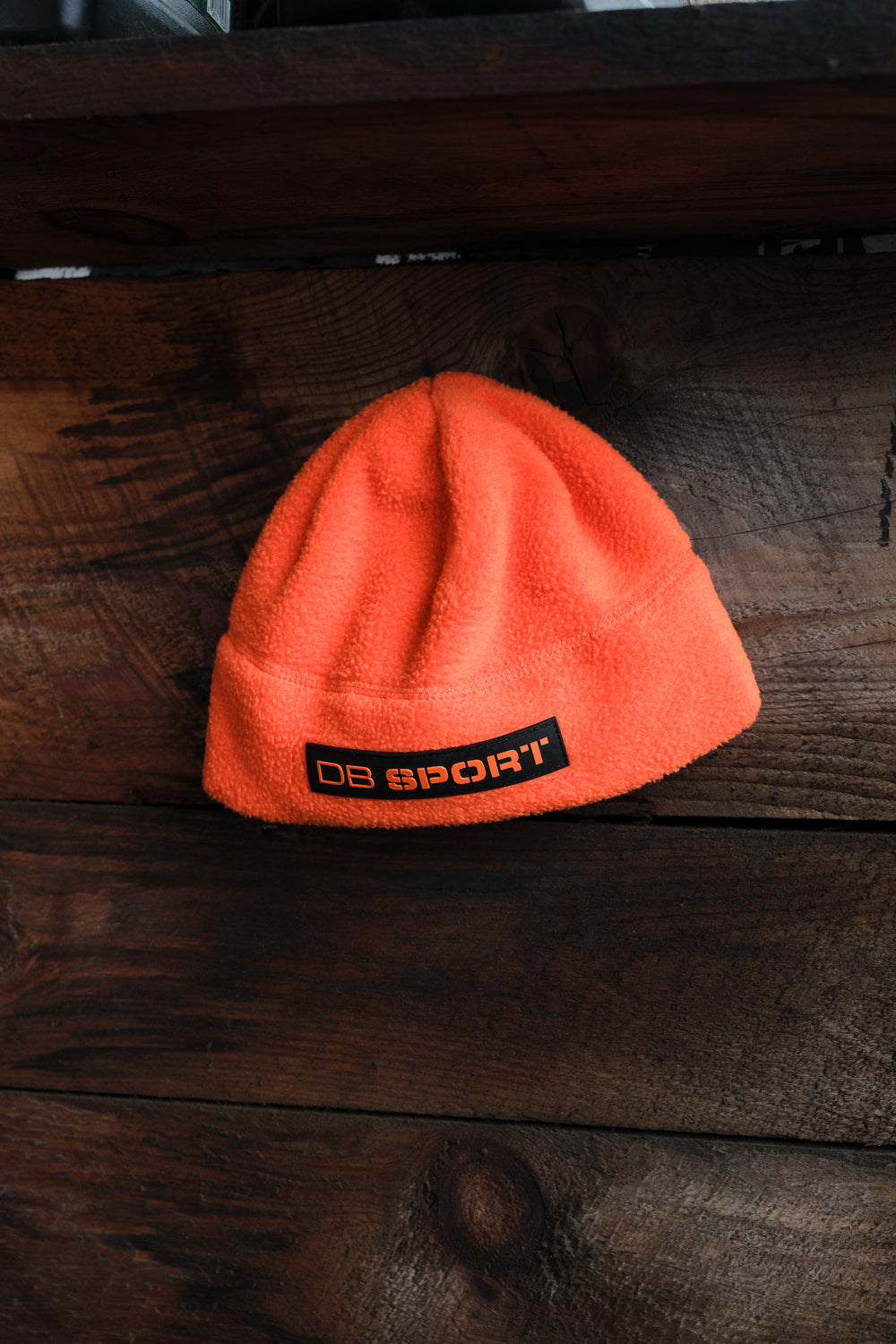 A bright orange fleece beanie hat with a black rectangular patch reading 'DB SPORT' in orange, resting on a dark rustic wooden surface.