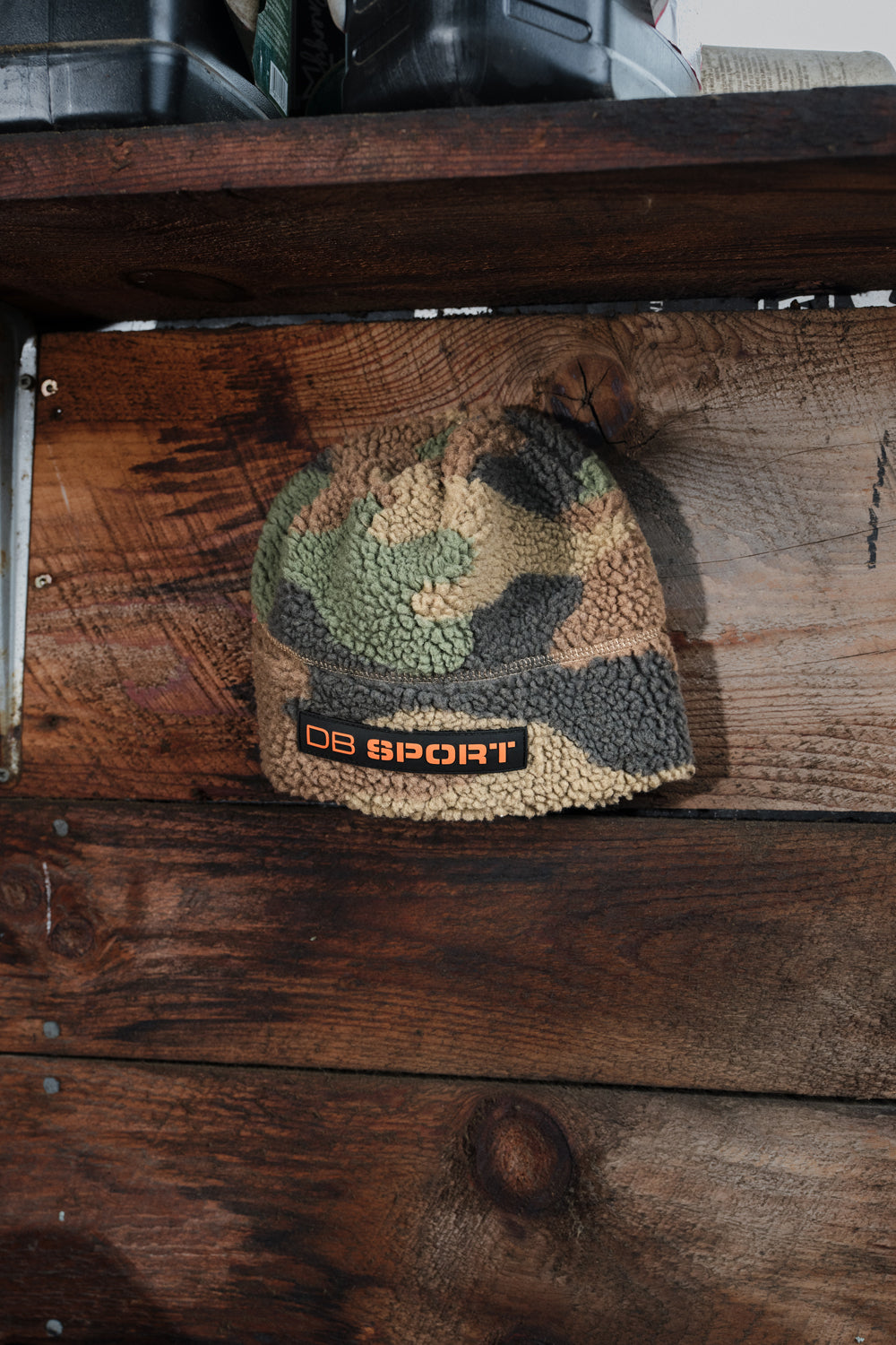 A fleece beanie hat with a camouflage pattern and an orange 'DB SPORT' logo patch, resting on a rustic wooden surface.