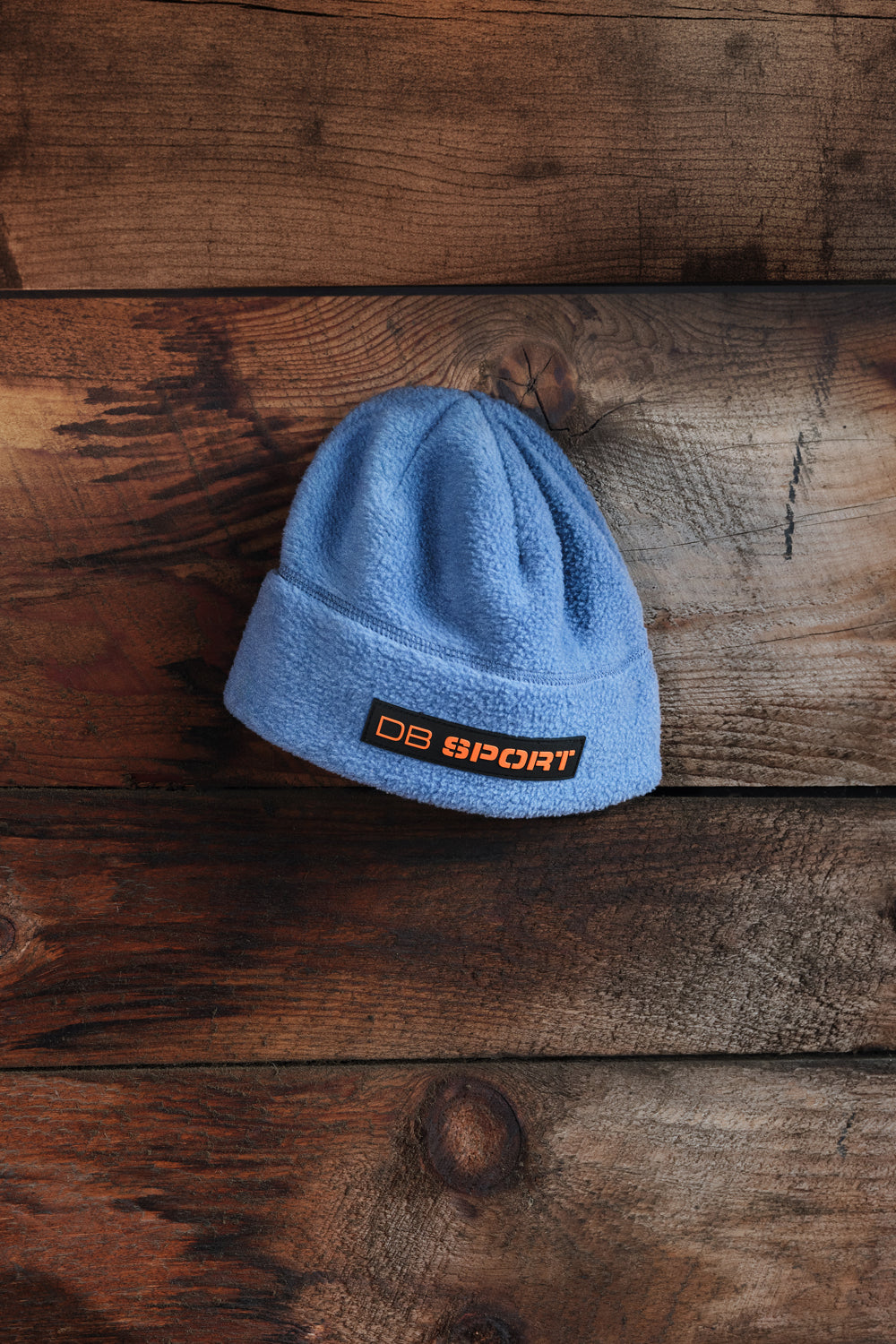 A light blue knit beanie hat with a black and orange 'DB SPORT' logo patch on the front cuff, displayed on a rustic dark brown wooden plank background.