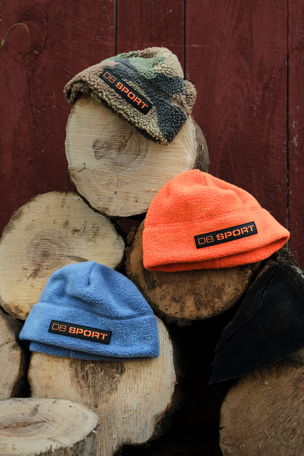 Three knit beanie hats displayed on stacked wood logs against a dark red wooden wall: one camouflage pattern, one bright orange, and one light blue. Each beanie has a black rectangular patch with the text 'DB SPORT' in orange.