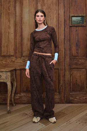 A woman stands in a rustic room with wooden paneling, wearing a matching dark brown camo patterned long-sleeve top and wide-leg pants, with light blue cuffs visible on her arms.
|M1