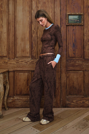 A woman stands in a rustic room with wooden paneling and flooring, wearing a matching brown camo patterned lounge set consisting of a long-sleeve top and wide-leg pants.
|M1