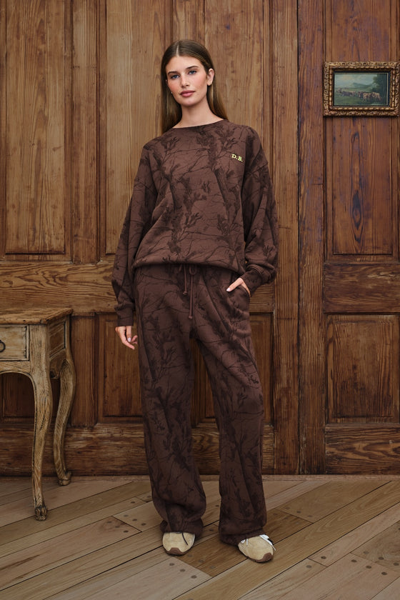 A woman stands in a rustic room with wooden paneling and a wooden floor, wearing a matching brown camo textured sweatshirt and sweatpants lounge set with beige slip-on shoes.
|M1