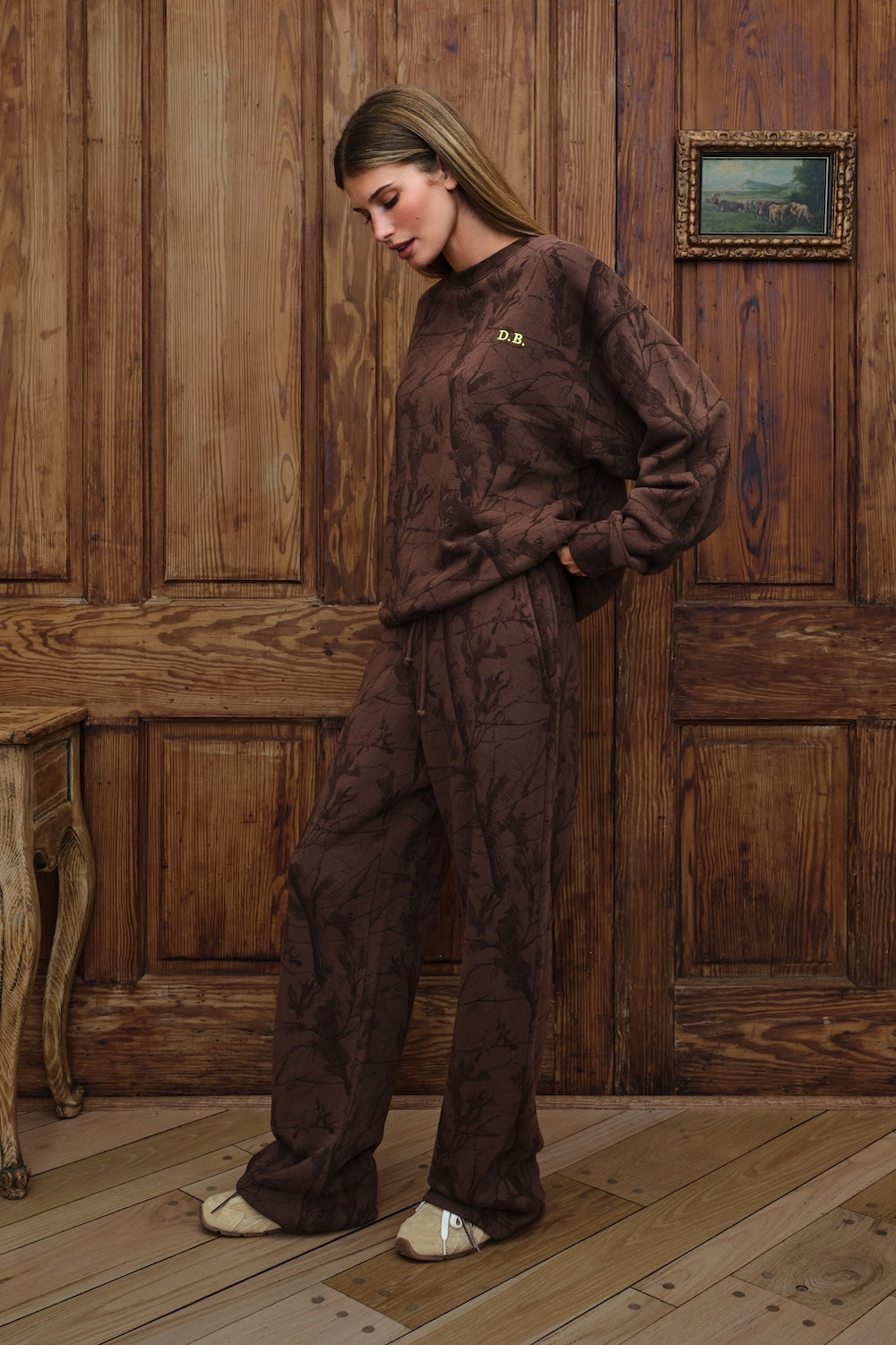 A woman wearing a matching dark brown patterned lounge set consisting of a sweatshirt and wide-leg pants, standing in front of a rustic wooden paneled wall.
|M1