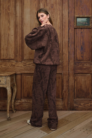 A woman wearing a matching brown camo patterned lounge set with a cropped long-sleeve top and wide-leg pants, standing in a rustic room with wooden paneling and a wooden floor.
|M1