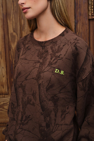 A close-up of a woman wearing a brown crewneck sweatshirt with a dark tree-branch camouflage pattern and a small yellow 'D.B.
|M1