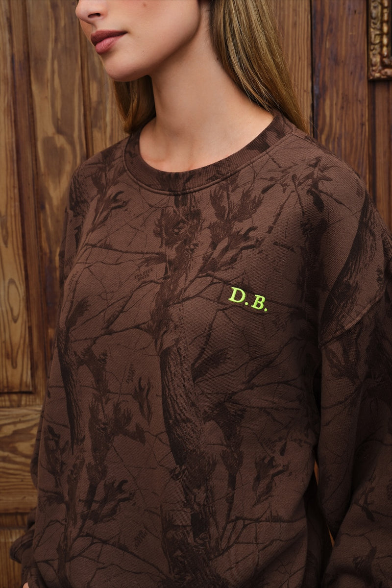 A close-up of a woman wearing a brown crewneck sweatshirt with a dark tree-branch camouflage pattern and a small yellow 'D.B.
|M1
