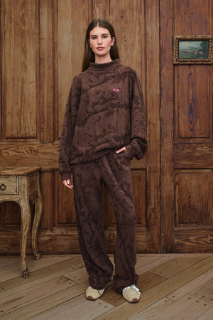 Model stands wearing brown camo rollneck sweatshirt with pink "D.B" monogram logo on the chest, in from of a rustic wood paneled wall. 
|M1