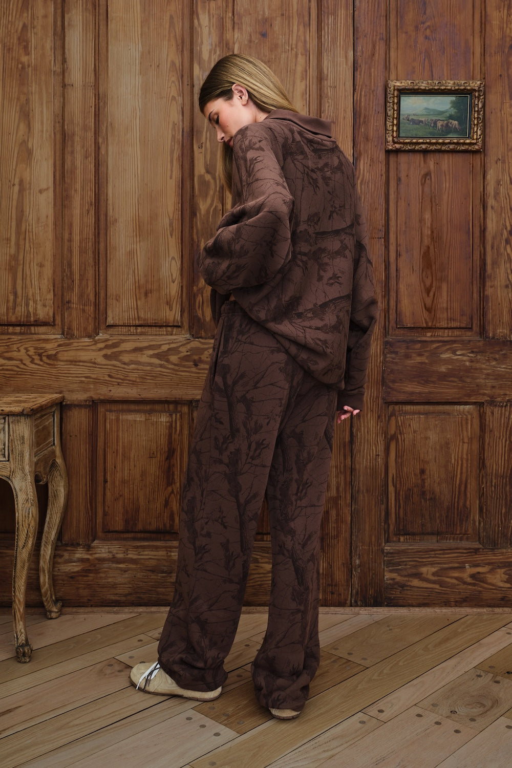 A woman stands in a room with wooden paneling and wood flooring, wearing a matching dark brown patterned lounge set consisting of a mockneck sweatshirt and wide-leg pants.
|M1