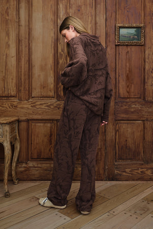 A woman stands in a room with wooden paneling and wood flooring, wearing a matching dark brown patterned lounge set consisting of a mockneck sweatshirt and wide-leg pants.
|M1