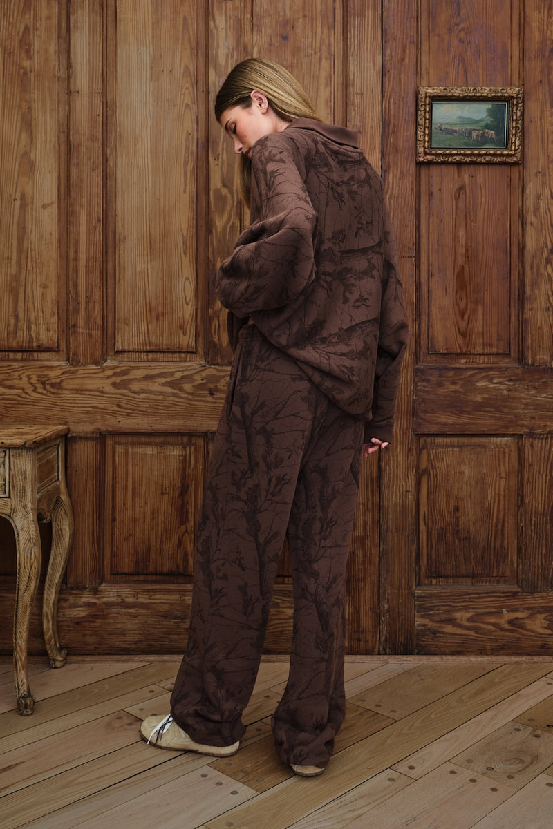A woman stands in a room with wooden paneling and wood flooring, wearing a matching dark brown patterned lounge set consisting of a mockneck sweatshirt and wide-leg pants.
|M1