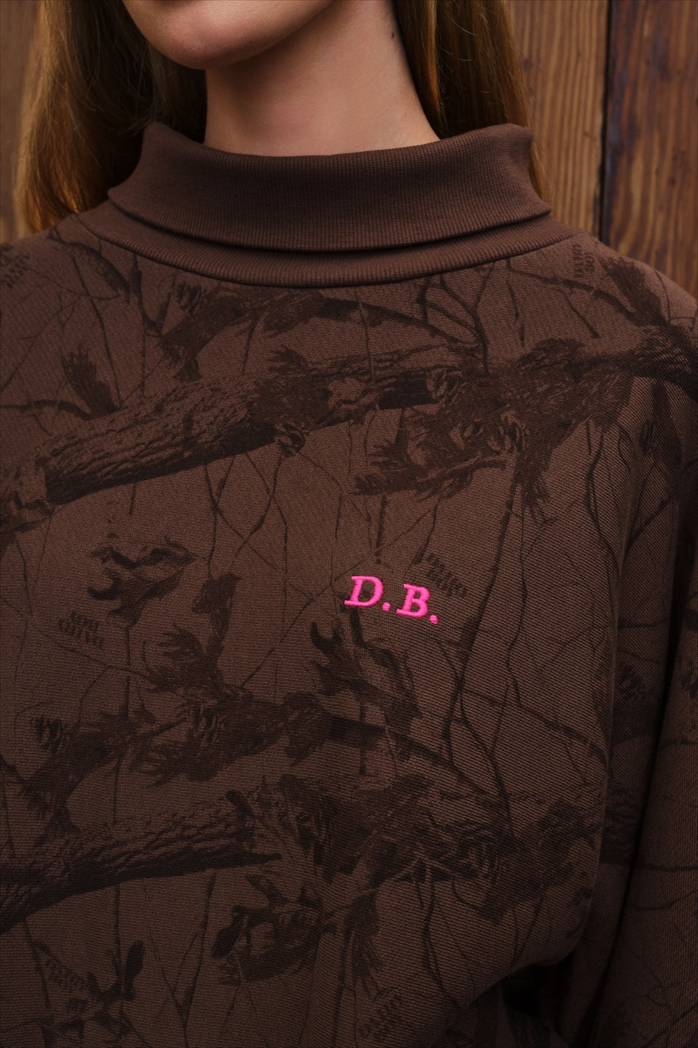Model stands wearing brown camo rollneck sweatshirt with pink "D.B" monogram logo on the chest, in from of a rustic wood paneled wall. 