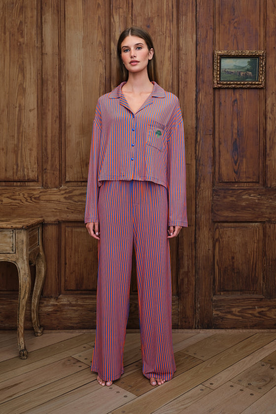 A woman stands in a rustic room with wooden paneling and flooring, wearing a matching two-piece pajama set with vertical orange and blue stripes. The set includes a long-sleeve button-up shirt and wide-leg pants.
|M1