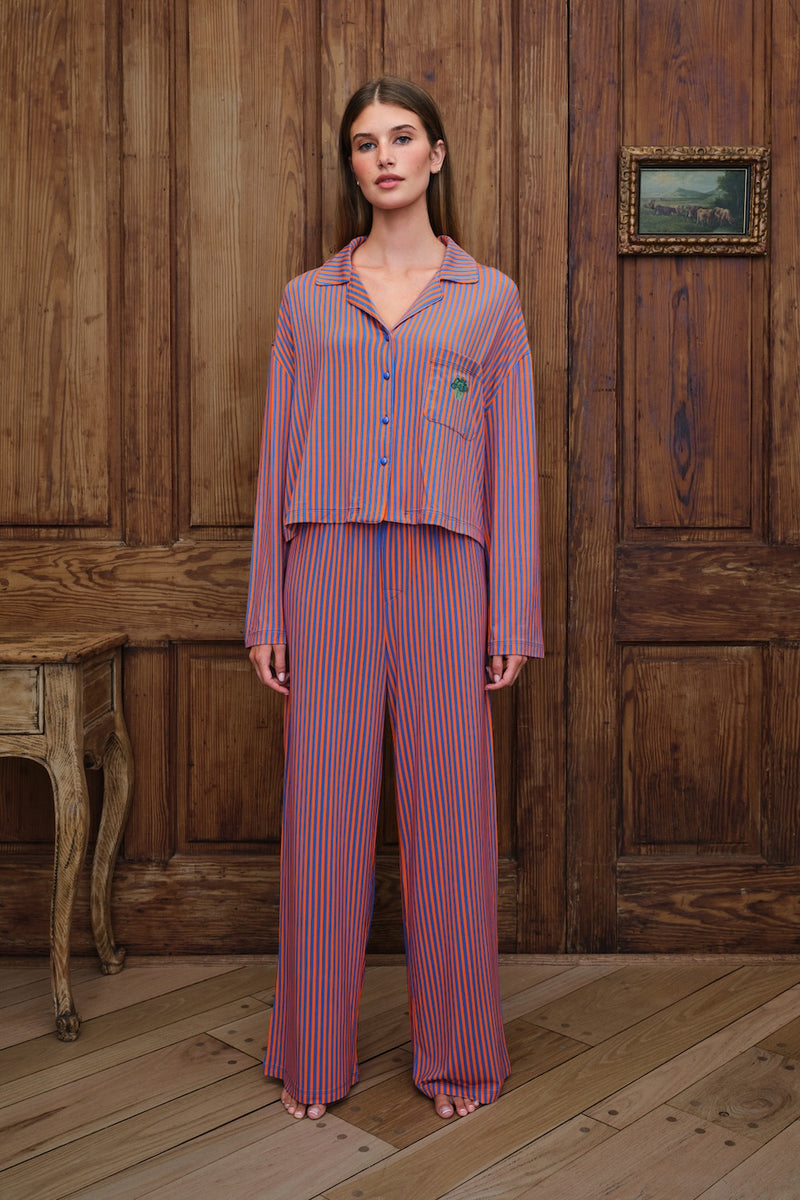 A woman stands in a rustic room with wooden paneling and flooring, wearing a matching two-piece pajama set with vertical orange and blue stripes. The set includes a long-sleeve button-up shirt and wide-leg pants.
|M1