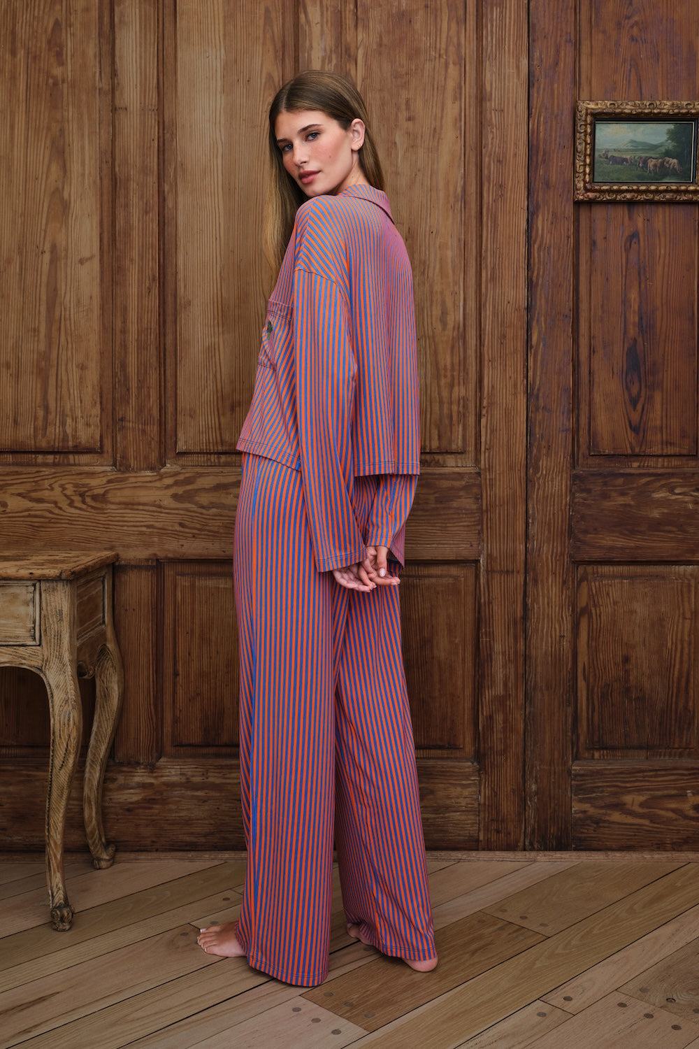 A woman stands barefoot in a rustic room with wooden paneling, wearing a matching pajama set with orange and blue vertical stripes. The set includes a long-sleeve button-up top and wide-leg pants.
|M1