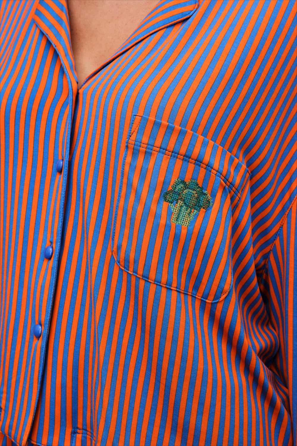 Close-up of a button-front shirt with narrow orange and blue vertical stripes and a small embroidered green mushroom on the chest pocket.