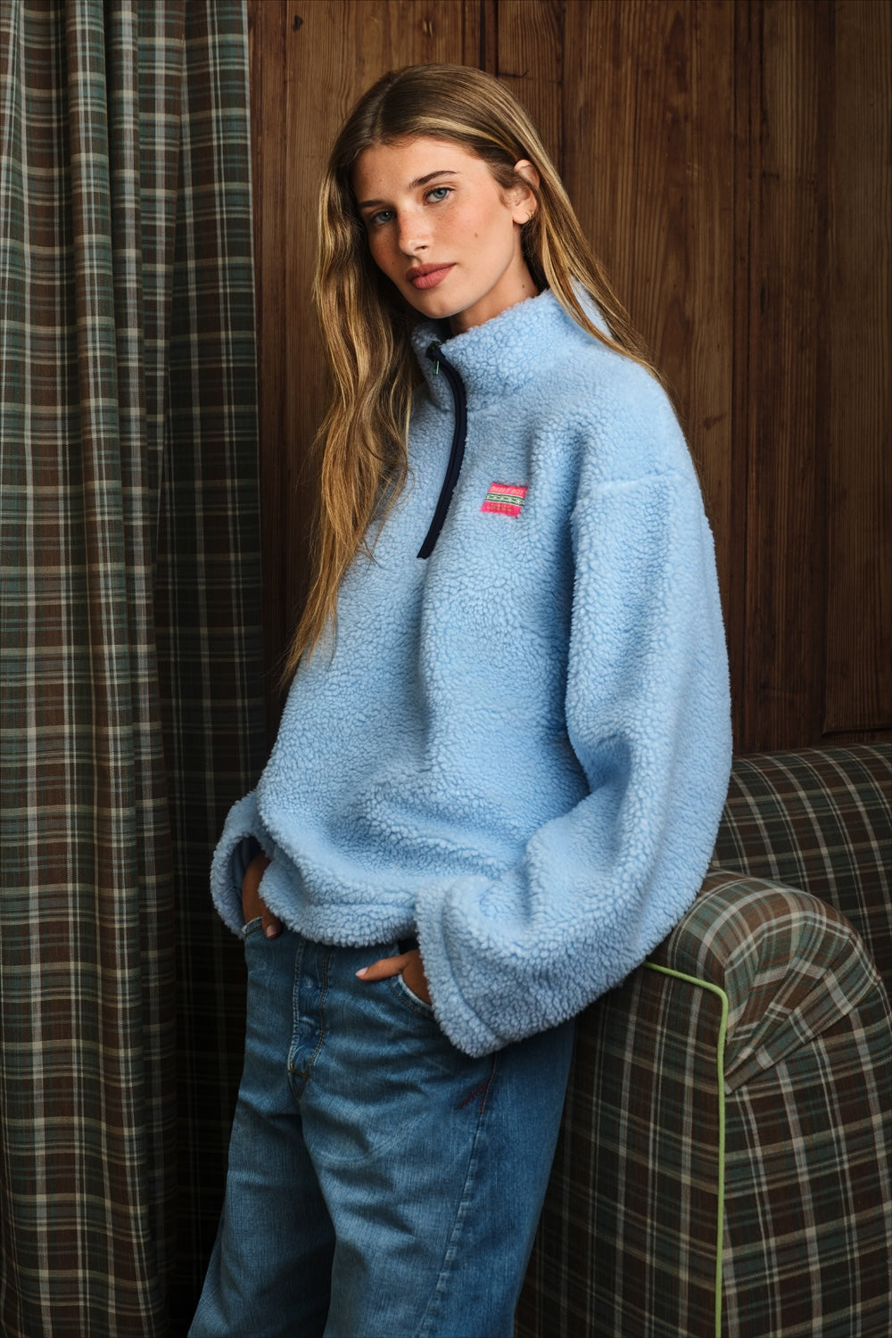 A woman wearing a light blue sherpa fleece quarter-zip pullover sweater with a small pink and white logo patch on the chest, paired with blue jeans, leaning against a plaid armchair in front of a wooden wall.
|M1