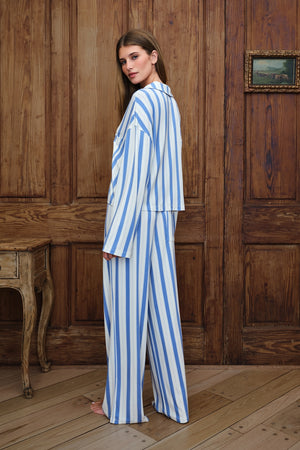 A woman wearing a blue and white vertically striped pajama set stands barefoot in a rustic room with wooden paneling and a wooden floor.
|M1