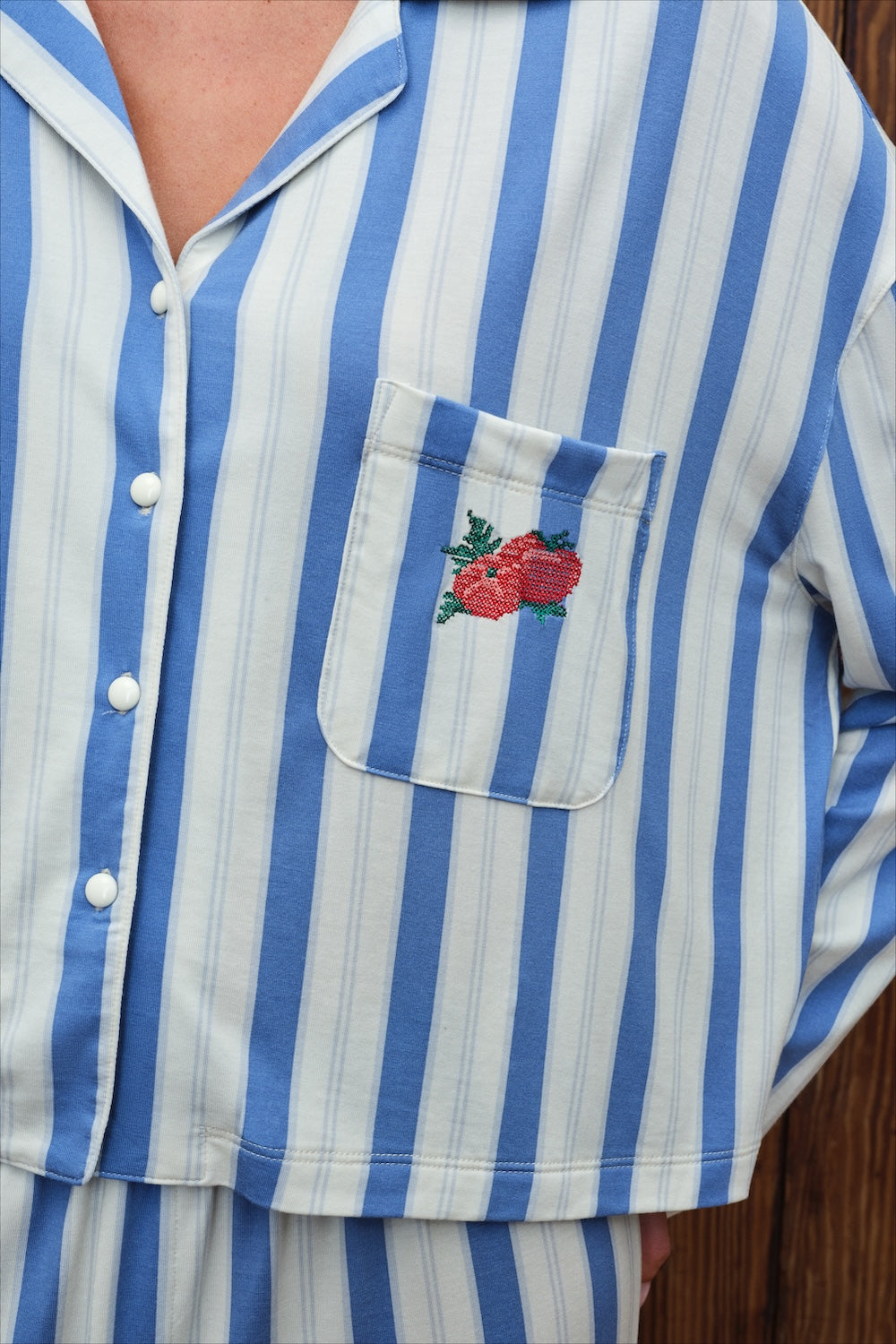 A close-up of a person wearing a blue and white vertically striped pajama shirt with a chest pocket featuring embroidered red tomatoes with green leaves.