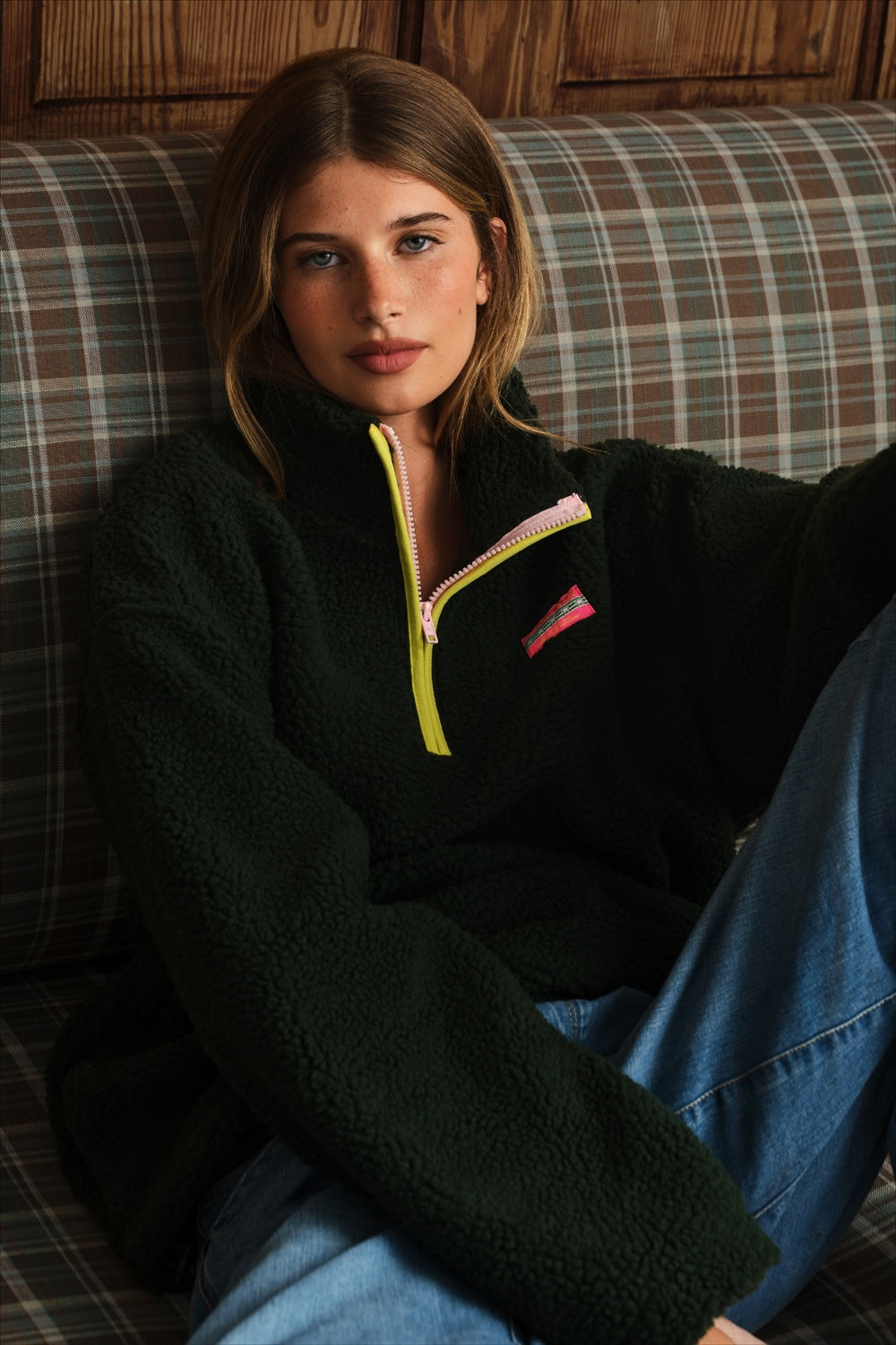 Model sitting on a plaid couch, wearing a dark green sherpa fleece pullover with a yellow zipper and pink logo patch on the chest.
|M1