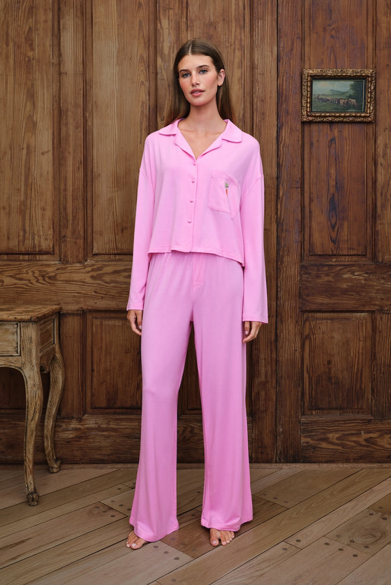 A woman stands barefoot in front of a rustic dark wood paneled wall, wearing a light pink two-piece pajama set consisting of a long-sleeve button-front top and matching wide-leg pants.
|M1