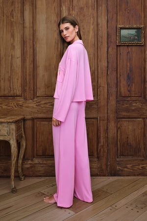 A woman wearing a light pink two-piece lounge set consisting of a long-sleeve top and matching wide-leg pants, standing in front of a rustic wooden paneled wall.
|M1