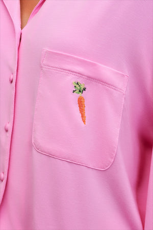 A close-up of a pink button-front shirt or pajama top with a chest pocket featuring an embroidered carrot motif with green leaves.