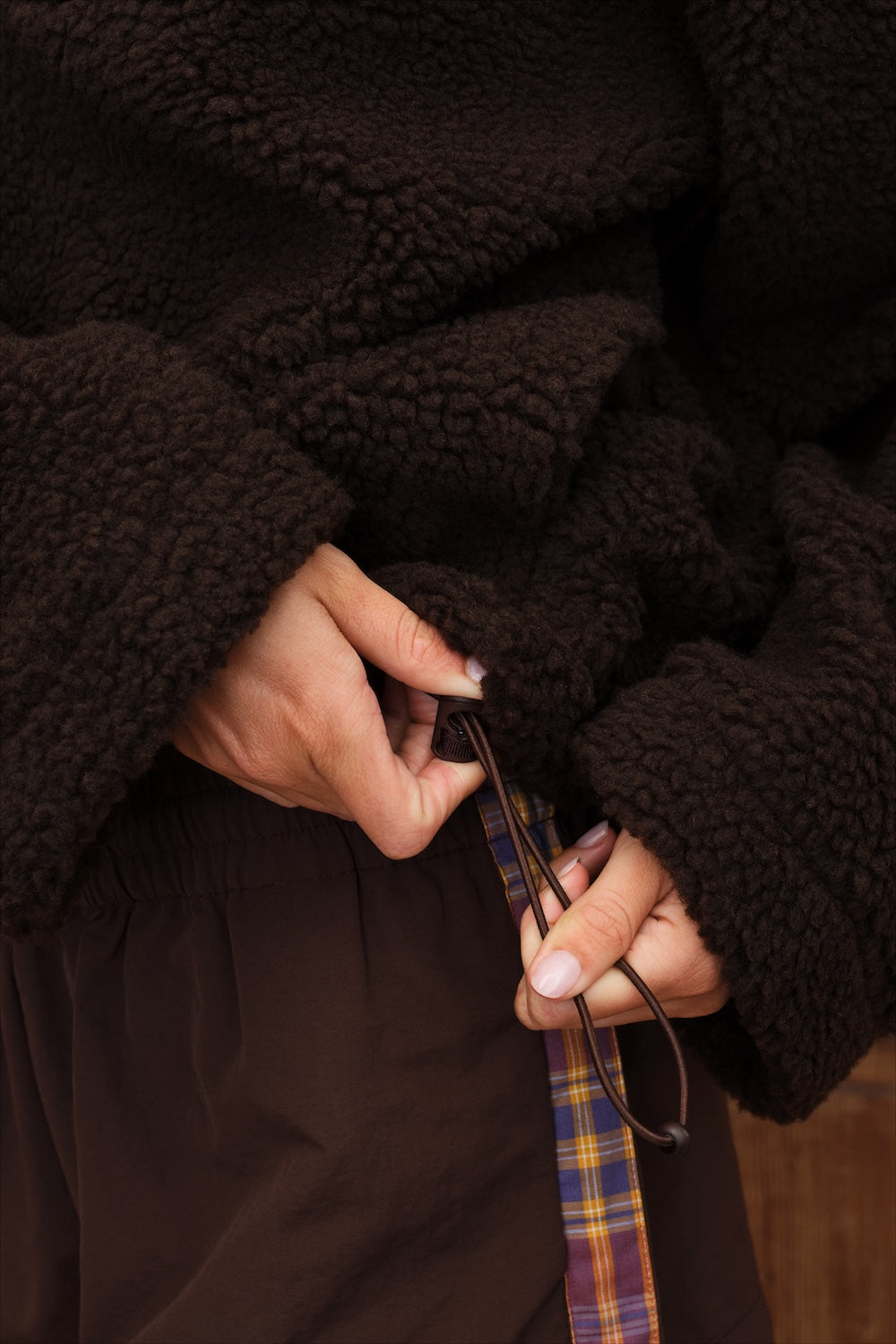 Close-up of a person wearing a dark brown sherpa fleece jacket and matching brown pants, adjusting a drawstring.