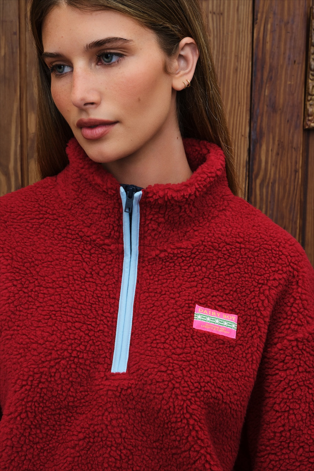 A close-up of a woman wearing a red sherpa fleece quarter-zip pullover with a light blue zipper and a pink and green logo patch on the chest, set against a rustic wooden background.
|M1