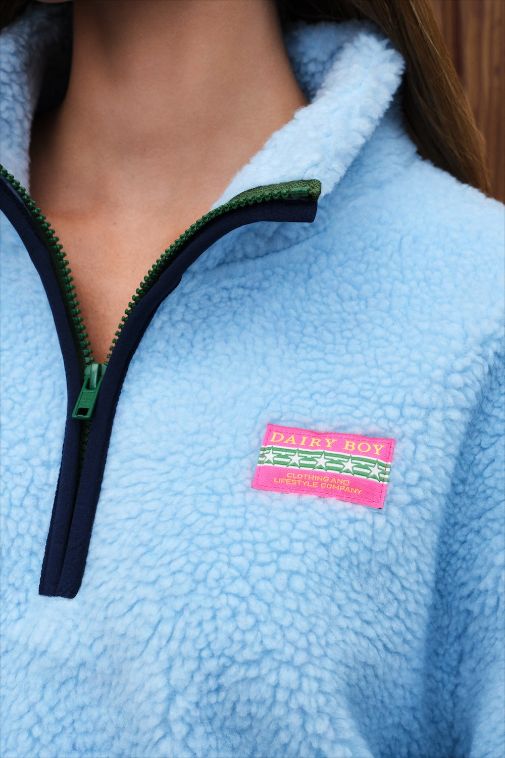 Close-up of a person wearing a light blue sherpa fleece quarter-zip pullover with a green zipper track and a pink 'DAIRY BOY' brand patch on the chest.
|M1